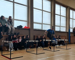 Uczennica pokonuje skok przez płotki, w tle widać innych uczniów, którzy siedzą jako publiczność.