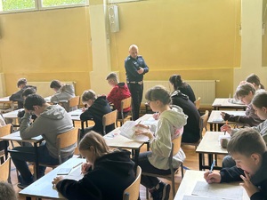 Sala gimnastyczna. Przy stolikach siedzą uczniowie, piszą test. Przy nich chodzi policjant i obserwuje jak piszą.