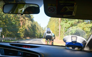 Zdjęcie zrobione z wnętrza radiowozu. Widać na desce rozdzielczej biała czapkę policyjną, a po drodze, przed radiowozem jedzie rowerzysta.