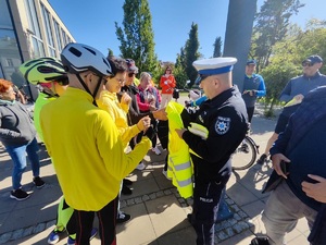 Umundurowany policjant, rozdaje uczestnikom rajdu rowerowego odblaskowe kamizelki i opaski. Wokół niego zebrała się grupa ludzi.