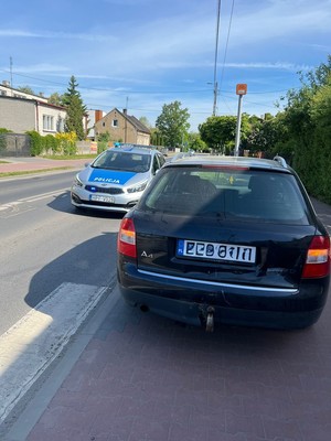 Na chodniku stoi tyłem samochód marki Audi, po jego lewej stronie, na drodze przy chodniku, stoi oznakowany radiowóz.