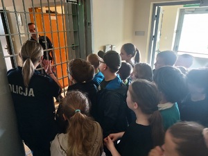 Policjantka pokazuje dzieciom pomieszczenie dla osób zatrzymanych, dzieci patrzą na pomieszczenia zza zamkniętych krat.