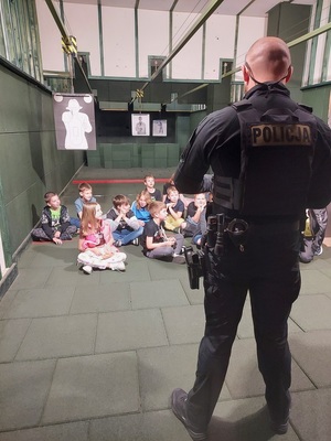 Dzieci siedzą na podłodze na strzelnicy. Przed nimi stoi policjant.
