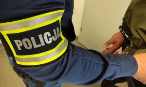 Policjant ubrany po cywilnemu, ma na prawym przedramieniu opaskę z napisem Policja. Przed nim stoi osoba zatrzymana, ma kajdanki założone na ręce trzymane z tyłu.