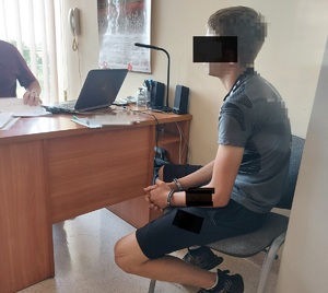 Po prawej stronie na krzesełku, siedzi osoba zatrzymana, ma kajdanki założone na ręce trzymane z przodu. Przed nim za biurkiem, przed komputerem siedzi policjant.