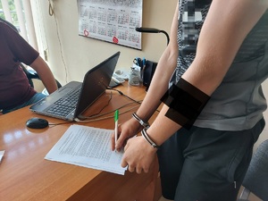 Osoba zatrzymana, ma kajdanki założone na ręce trzymane z przodu, trzyma w ręku długopis, podpisuje dokument leżący na biurku.