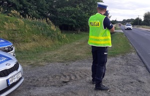 Umundurowany policjant stoi tyłem, patrzy na drogę i jadące samochody przez urządzenie do pomiaru prędkości. Po jego lewej stronie stoi radiowóz policyjny, po prawej jest droga.