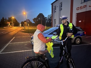 Policjant ruchu drogowego rozmawiający z rowerzystką, który wręcza kobiecie kamizelkę odblaskową.