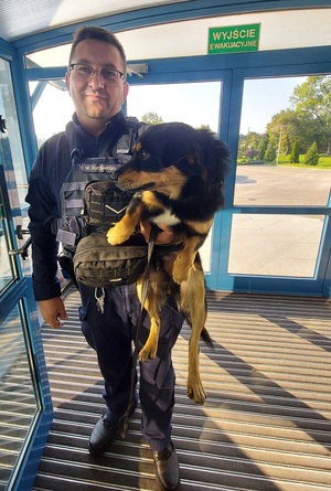 Policjant stoi w drzwiach, trzyma pod pachą psa.