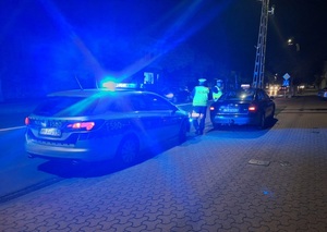 Jest ciemno, widać światła radiowozu, policjanci stoją przy taksówce.