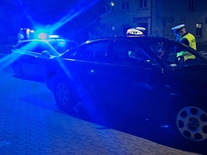 Jest ciemno, widać światła radiowozu, policjanci stoją przy taksówce.