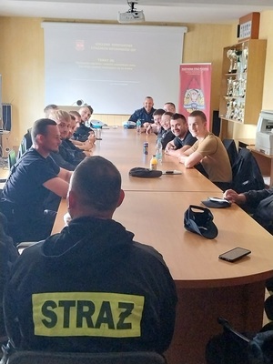 W sali, przy długim stole, siedzą z obu stron strażacy, w szczycie stołu, na wprost siedzi policjant ruchu drogowego i przeprowadza szkolenie.