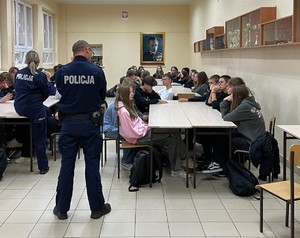 Sala lekcyjna, wzdłuż długiego stołu siedzą po obu stronach uczniowie, w szczycie stołu tyłem do zdjęcia stoi policjant i policjantka, rozmawiają z młodzieżą.