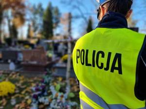 Tyłem stoi policjant w kamizelce odblaskowej z napisem Policja, w tle widać groby.