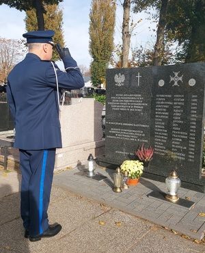 Przed tablicą, tyłem do zdjęcia, stoi w galowym umundurowaniu Komendant Powiatowy Policji w Zduńskiej Woli. Salutuje.