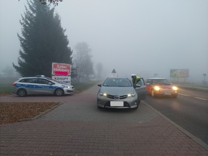 Jest mgła, po lewej stronie stoi radiowóz, po środku stoi samochód osobowy, po jego prawej stronie stoi policjant i rozmawia z kierowcą, a za nim jest ulica którą jadą inne samochody.