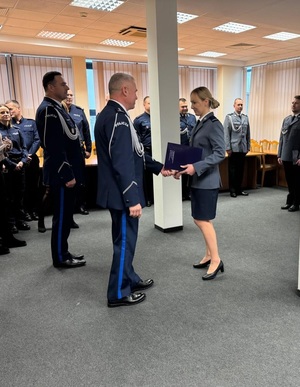 Komendant gratuluje stojącemu przed nim policjantowi, za komendantem stoi jego zastępca, a dalej kilka osób, które są publicznością.