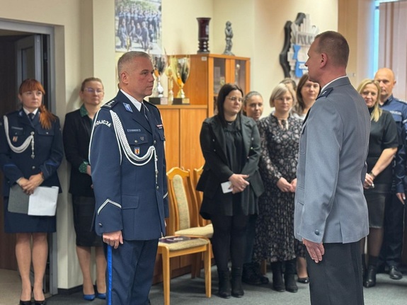 Policjant składa meldunek komendantowi. W tle widać stojących kilka osób bez mundurów, które są publicznością.