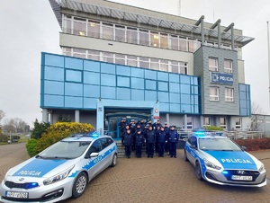 Przed budynkiem komendy stoją dwa radiowozy. Między autami, na schodach, stoją umundurowani policjanci. Salutują.