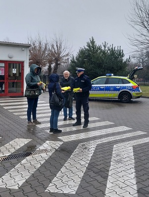 Policjant wręcza osobie kamizelkę odblaskową. W tle radiowóz.