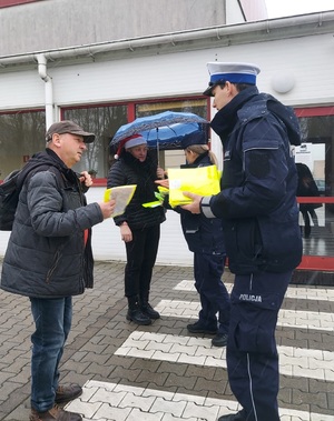 Policjant wręcza osobie kamizelkę odblaskową. Dalej stoją policjantka i kobieta, która trzyma rozłożony nad głową parasol.