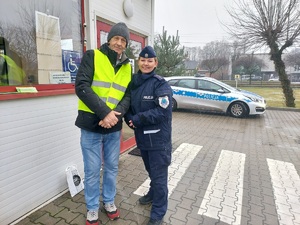 Mężczyzna ma założoną na kurtkę odblaskową kamizelkę. Obok niego stoi uśmiechnięta policjanta. Pozują razem do zdjęcia.