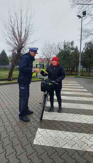 Policjant wręcza kamizelkę odblaskową kobiecie, która prowadzi rower.