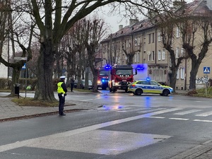 Skrzyżowanie. Stoi radiowóz, a także samochody straży pożarnej. Policjant stoi w pobliżu, obserwuje otoczenie. W tle bloki mieszkalne.