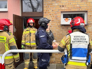 Policjant rozpoznania minersko-pirotechnicznego stoi pod budynkiem, wokół niego stoją strażacy.