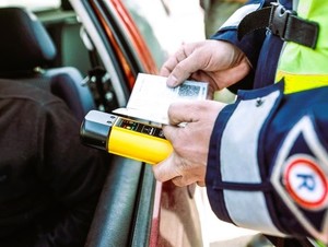 Przy otwartym oknie samochodu od strony kierowcy stoi umundurowany policjant, trzyma w ręku dokument, a także urządzenie do pomiaru stanu trzeźwości.