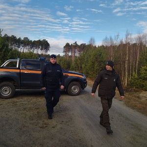 Policjant i strażnik leśny idą drogą, za nimi stoi samochód służbowy Straży Leśnej.