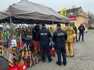 Policjant wspólnie ze strażakiem i strażnikiem miejskim kontrolujący stoisko z wyrobami pirotechnicznymi.