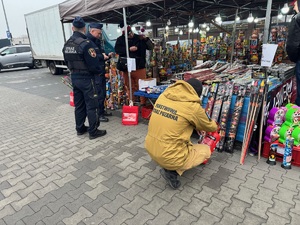 Umundurowany policjant rozmawiający ze sprzedawcą fajerwerków i strażak sprawdzający wyroby przeciwpożarowe na stoiku.