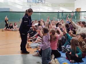 Hala sportowa w szkole. Policjantka rozdaje uczniom ulotki dotyczące konkursu &quot;Śnieżny dekalog&quot;. Uczniowie siedzą na materacach na podłodze i zgłaszają się, by policjanta dała im ulotki.