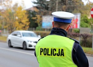 Umundurowany policjant, stoi przy drodze, patrzy na jadące samochody.