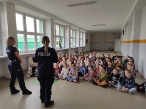 Korytarz szkolny. Dzieci siedzą na podłodze, na wprost nich stoją dwie policjantki, mówią do dzieci.