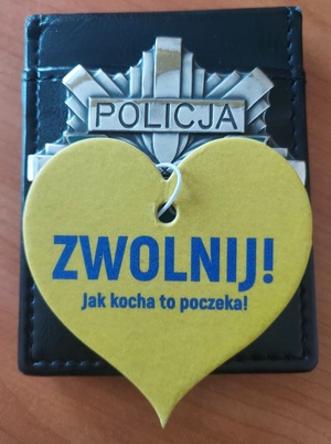 Zdjęcie odznaki policyjnej, na której leży serduszko zapachowe z napisem &quot;Zwolnij&quot; Jak kocha to poczeka!&quot;