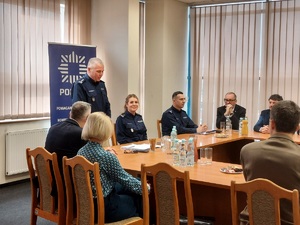 Aula w komendzie, przy stołach, na krzesłach siedzą umundurowani policjanci i goście ubrani po cywilnemu oraz reprezentacji zaprzyjaźnionych służb. Jeden z policjantów stoi i mówi do zebranych.