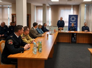 Aula w komendzie, przy stołach, na krzesłach siedzą umundurowani policjanci i goście ubrani po cywilnemu oraz reprezentacji zaprzyjaźnionych służb. Jeden z policjantów stoi i mówi do zebranych.