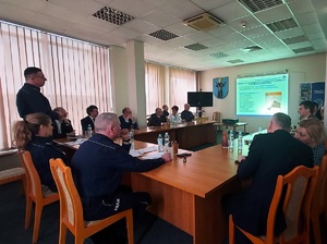 Aula w komendzie, przy stołach, na krzesłach siedzą umundurowani policjanci i goście ubrani po cywilnemu oraz reprezentacji zaprzyjaźnionych służb. Jeden z policjantów stoi i mówi do zebranych. Na ścianie auli widać wyświetlany z rzutnika obraz.