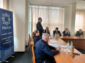 Aula w komendzie, przy stołach, na krzesłach siedzą umundurowani policjanci i goście ubrani po cywilnemu oraz reprezentacji zaprzyjaźnionych służb. Jeden z policjantów stoi i mówi do zebranych.