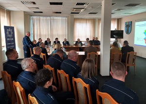 Aula w komendzie, przy stołach, na krzesłach siedzą umundurowani policjanci i goście ubrani po cywilnemu oraz reprezentacji zaprzyjaźnionych służb. Jeden z policjantów stoi i mówi do zebranych. Na ścianie auli widać wyświetlany z rzutnika obraz. Na pierwszym planie zdjęcia widać siedzących policjantów - kadrę kierowniczą jednostki.