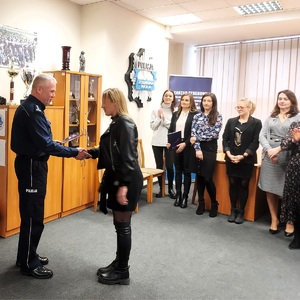 Aula w jednostce. Komendant gratuluje kobiecie. W tle stoją inne osoby.