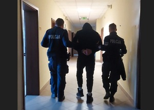 Zatrzymany w kapturze na głowie, idzie korytarzem, jest prowadzony przez dwóch umundurowanych policjantów.