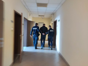 Zatrzymany idzie korytarzem, jest prowadzony przez dwóch umundurowanych policjantów.