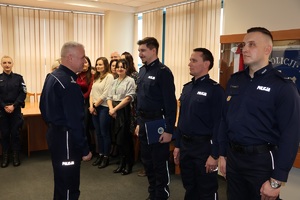 Komendant stoi przed trzema mężczyznami - umundurowanymi policjantami. W tle widać inne stojące osoby.