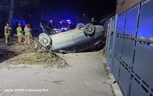 Noc, samochód leży na dachu, widać wokół niego połamane drzewo. W tle idzie strażak.