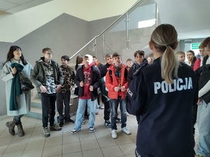 Policjantka stoi tyłem do zdjęcia, a przodem stroją dzieci, patrzą się na mówiąca do nich policjantkę.