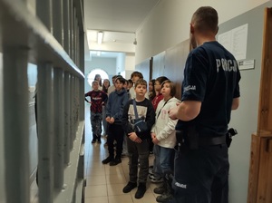 Policjant stoi tyłem do zdjęcia, przodem do dzieci. Są w pomieszczeniu dla osób zatrzymanych.