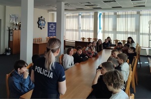 Aula komendy. Dzieci siedzą na krzesełkach wzdłuż stołu. W szczycie stołu stoi policjantka i mówi do zebranych.